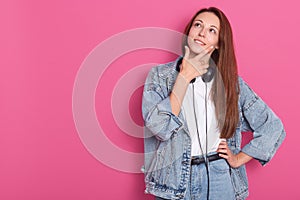 Image of cute playful young woman smiling and thinking, keeps hand under chin, looking up, wearing denim jacket, jeans and white t