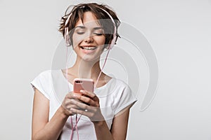 Image of cute woman in basic t-shirt holding smartphone while listening to music with headphones