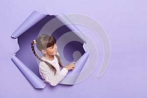 Image of dark haired little girl wearing striped shirt posing in torn purple paper hole, standing looking away at copy space for