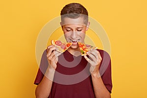 Image of delighted hungry young man eating two slices of pizza, being fond of junk food, wearing red t shirt, standing isolated