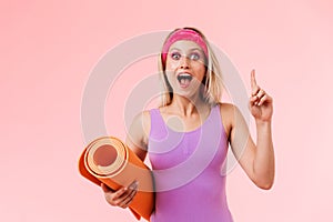 Image of delighted sporty woman holding mat and pointing finger upward