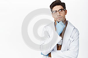 Image of doctor in glasses and medical gloves thinking, looking up with thoughtful face, standing over white background