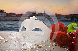 Image of elegant venetian mask and red roses over wooden table in front of blurry Venice background.