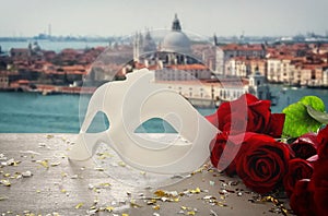 Image of elegant venetian mask and red roses over wooden table in front of blurry Venice background.