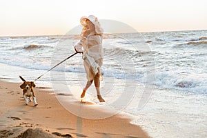 Image of european woman 20s in summer straw hat, running by seaside with her dog