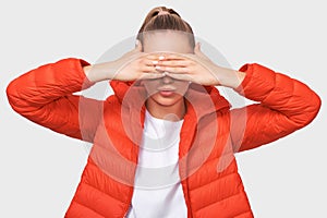 Image of European young woman in casual red jacket, covering her eyes with both hands, waiting for a surprise for her birthday.