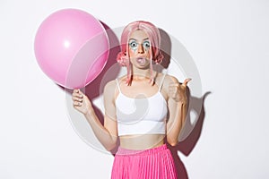 Image of excited attractive girl blowing bubble gum, showing thumbs-up in approval, standing with cute pink balloon