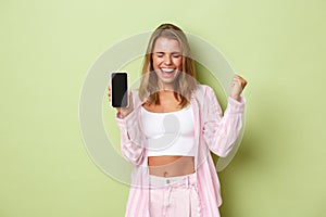 Image of excited blond girl winning something, showing app achievement on mobile phone screen, saying yes and triumphing