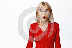 Image of excited blond stylish girl looking away at copy space, pucker lips from surprise, amazed by smth, standing in