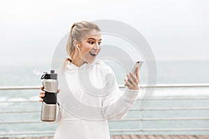 Image of excited blonde woman using earpods and cellphone while drinking water from bottle in boardwalk