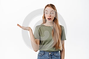 Image of excited girl looks at her hand holding copyspace with interest, display item on open palm, standing against