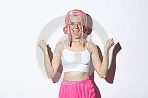 Image of excited successful girl in pink wig, celebrating something, making fist pump and smiling satisfied, enjoying