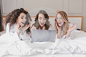 Image of excited surprised women 20s wearing white housecoat lying in luxury bedroom at home, and looking at silver laptop during photo
