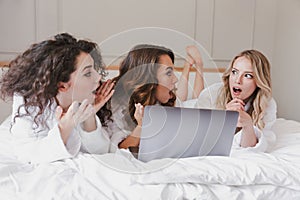 Image of excited surprised women 20s wearing white housecoat lying in luxury bedroom at home, and using silver laptop during hen photo