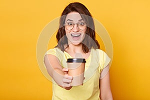 Image of excited young beautiful female handing takeaway coffee, stretching hand with hot beverage to her friend, having