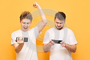 Image of excited young two men playing together and competing in video games on smartphones isolated over yellow background