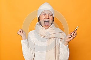 Image of extremely happy overjoyed woman wearing warm jumper, hat and scarf, holding mobile phone in hands, clenched fists,