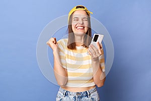 Image of extremely happy smiling teen girl wearing baseball cap and casual T-shirt, using cell phone, reading message with
