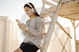 Image of fair-skinned young teenager girl wearing headphones picks song from playlist on her phone.