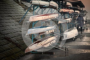 Multiple rowing shells, including sweep oar and coxed four boats, rest on racks by the riverside at sunset. These competitive photo