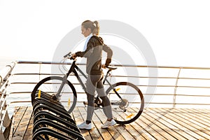 Image of fitness woman standing with bicycle on boardwalk, during sunrise over sea background