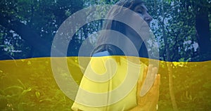 Image of flag of ukraine over caucasian woman praying outdoors