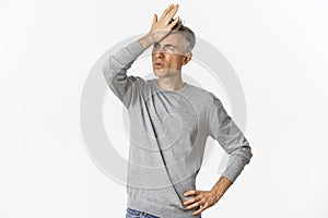Image of frustrated and distressed middle-aged man, making facepalm and exhaling disappointed, standing over white