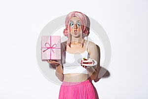 Image of funny birthday girl making silly faces, holding b-day cake and wrapped gift, standing over white background