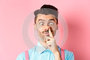 Image of funny caucasian guy picking nose and squint eyes, making dumb face, standing on pink background