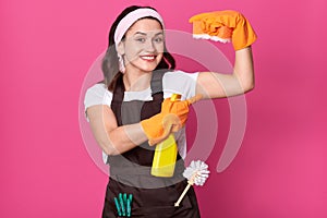 Image of funny charming young Caucasian housewife wearing headband, orange rubber gloves and brown apron, holding sponges and