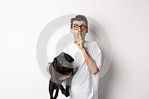 Image of funny young man holding cute black pug dog and shushing at camera, showing hush sign and staring at camera