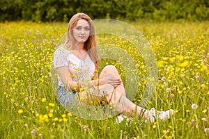 Image of girl sitting on grass in park or meadow, looking directly at camera, wearing casual white t shirt and denim skirt, being