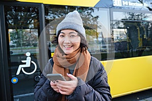 Image of girl student waiting for public transport, checks schedule on smartphone app, stands near city bus