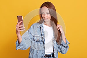 Image of girl in white t-shirt and demin jacket taking selfie  over yellow background, holding smartphone in hands,