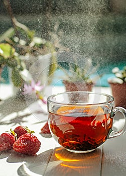 Image of glass cup of tea, red strawberries, flowers in pots on a light wooden table.