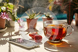Image of glass cup of tea, red strawberries, smartphone and flowers in pots on a light wooden table.
