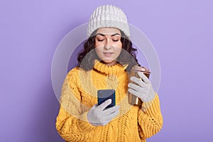 Image of good looking adorable attentive female wearing grey hat and gloves, holding smartphone and thermo mug with hot drink in