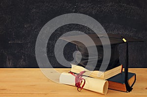 Image of graduation black hat over old books next to graduation on wooden desk. Education and back to school concept