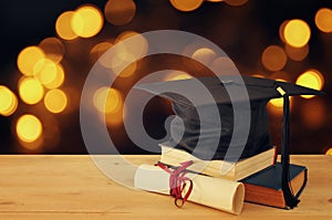 Image of graduation black hat over old books next to graduation on wooden desk. Education and back to school concept.