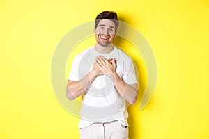 Image of grateful handsome guy in white t-shirt, holding hands on heart and smiling pleased, express gratitude, thanking