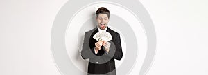 Image of handsome bearded guy in black suit, looking at money with excitement, standing over white background