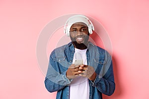 Image of handsome Black guy in headphones, listening music and using mobile phone, smiling at camera, pink background