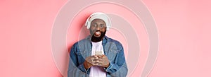 Image of handsome Black guy in headphones, listening music and using mobile phone, smiling at camera, pink background