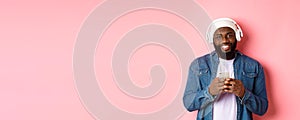 Image of handsome Black guy in headphones, listening music and using mobile phone, smiling at camera, pink background