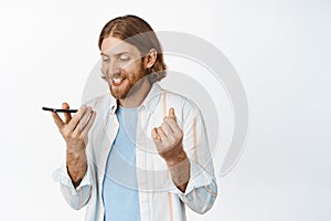 Image of handsome blond smiling man record voice message, holding mobile phone and talk on speakerphone, translate his