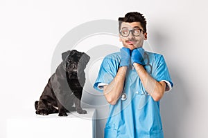 Image of handsome male doctor veterinarian looking at cute black pug dog sitting on table, admiring puppy cuteness