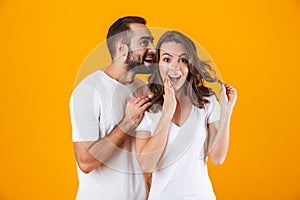 Image of handsome man whispering secret or interesting gossip to woman in her ear, isolated over yellow background