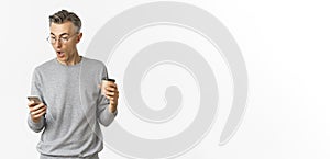 Image of handsome middle-aged man in grey sweater and glasses, drinking coffee and looking amazed at mobile phone screen