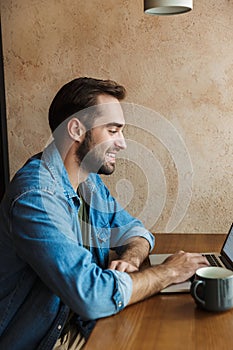 Image of handsome positive man smiling and using laptop while sitting at desk in cafe indoors