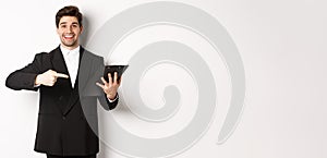 Image of handsome smiling businessman in black suit, pointing finger at clipboard with documents, standing against white